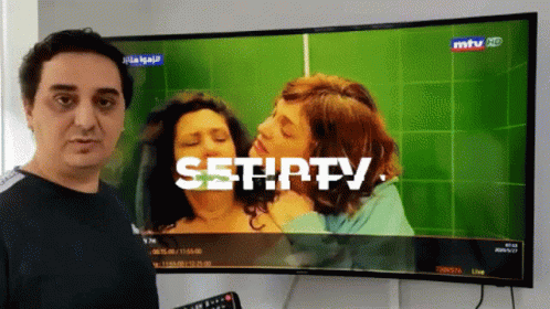 A man is standing in front of a television displaying a scene with two women embracing; the screen has the text "SETHFY" overlaid on it.